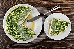 Knife on dish with section of omelet, slice of omelet with parsley and green peas in plate on wooden table. Top view