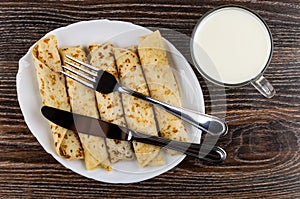 Knife and fork on white dish with pancake rolls, cup of milk on table. Top view