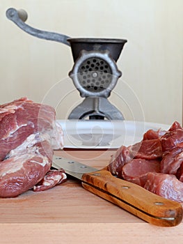 Knife and meat for mince on a wooden cutting board. Behind them are a vintage manual meat grinder and a white plate on a beige bac