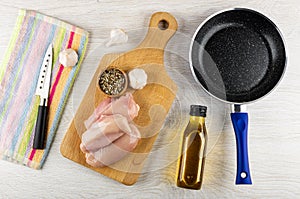 Knife on napkin, raw chicken breast, garlic, bowl with condiment on cutting board, bottle of vegetable oil, frying pan on wooden