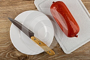 Knife on plate, sausage on cutting board on wooden table. Top view