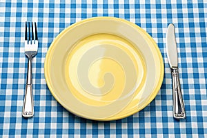 Knife, yellow plate, and fork on blue checked tablecloth