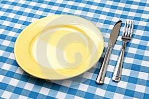 Knife, yellow plate, and fork on blue checked tablecloth