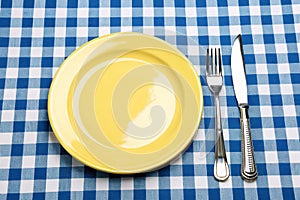 Knife, yellow plate, and fork on blue checked tablecloth