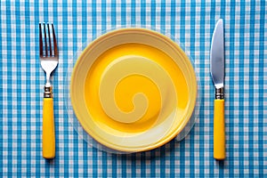 Knife, yellow plate, and fork on blue checked tablecloth