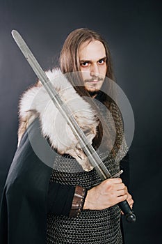 Knight in chain mail and with a fur collar on a black cloak on a gray background. Portrait of a Viking man with long hair and a