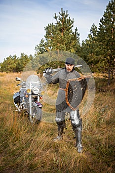 knight in chainmail with a shield and a sword in his hands stands against the backdrop of a motorcycle and a forest