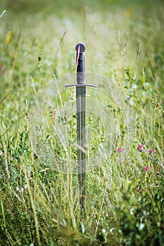 Knight`s sword thrust into the ground. In the field against the background of the forest and the blue sky.