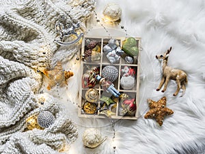 Knitted plaid, box of christmas toys, light garland on fluffy carpet, top view. Christmas background