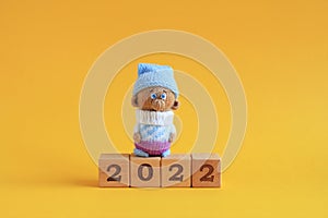 A knitted toy gnome stands on wooden cubes with numbers stacked up to number 2022 on a yellow background. Closeup