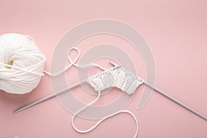 Knitting project in progress. White knitting wool and knitting needles on pastel pink background. Female hobby knitting