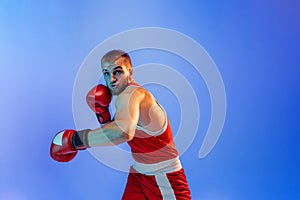 Knockout punch. Male boxer in red uniform and boxing gloves training  on blue background in neon. Strength