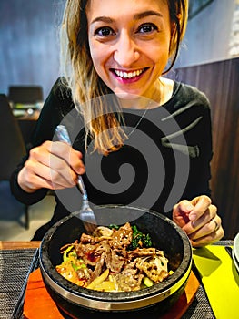 Korean bibimbap served in hot stone dolsot pot bowl eaten by caucasian smiling woman dining in korean ethnic restaurant