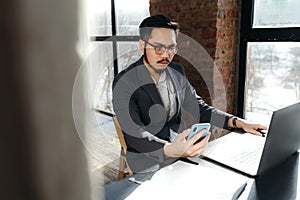 Korean businessman with glasses working in an office behind a laptop while holding a phone in his hand