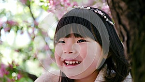 Korean girl in a white light fur coat and a headband sitting on a tree branch in a garden with cherry blossoms