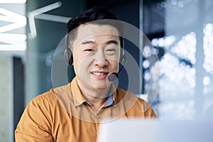 Korean man in a good mood office worker in a headset sits at a desk in front of a laptop in a modern office, an Asian