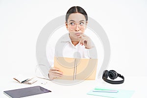 Korean woman hides her face behind notebook, peeks at camera, sits at desk with headphones and tablet, white background