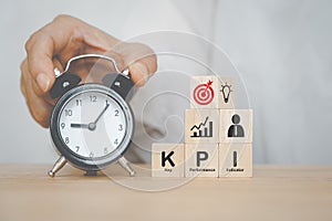 KPI, Key Performance Indicator, on cubes and business icon above with  hand holding black analog alarm clock