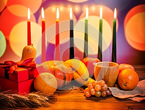 Kwanzaa festive concept with seven candles red, black and green, gift box, pumpkin, bowl and fruit on light blur bokeh background