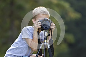 KYIV, UKRAINE - August 25, 2018: Young cute blond child boy taking picture with tripod camera on blurred green copy space