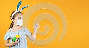 A l girl in a medical mask for coronavirus on her face, on her head with bunny ears, holds Easter eggs in her hands, points to an