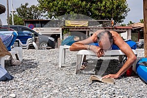 Old man wearing glasses reading a book lying on a sunbed on the beach