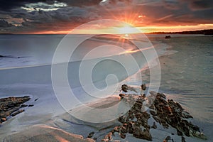 La Pelosa beach in Sardinia at sunrise with a sunburst and rocks in the foreground