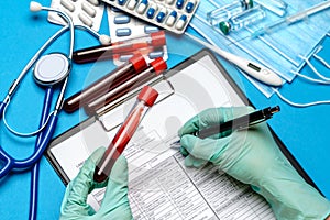 Lab technician assistant or doctor wearing rubber or latex gloves holding blood test tube over clipboard with blank form