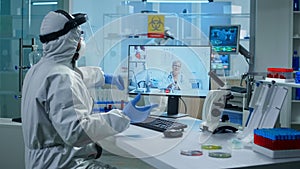 Lab technician in ppe suit holding test tube and talking on video call with doctor