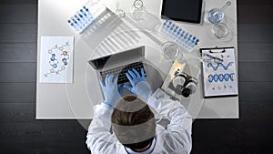 Lab worker viewing samples under microscope and typing results on laptop topview