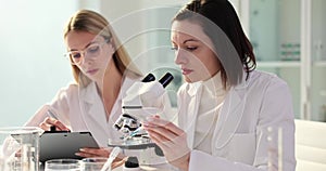 Laboratory assistant of biologist looks into microscope liquid drips and colleague writes medical report in laboratory