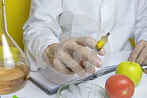 Laboratory assistant in a white robe nitrates colitis and GMOs in a tomato and an apple are lying on a table on a yellow