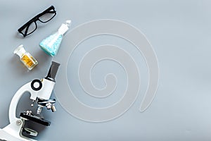 Laboratory desk with glasses, pills in test tube and microscope on gray background top view space for text