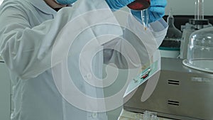 A laboratory technician in a white coat and protective gloves pours liquid into a chemical dish.