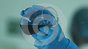 A laboratory technician in a white coat and protective gloves pours liquid into a chemical dish.