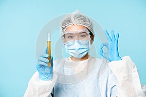 Laboratory worker, nurse in personal protective equipment, showing sample in test tube and okay sign, testing patient