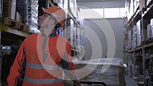 Laborer in uniform pulling trolley with boxes between rows of racks on warehouse