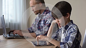 Lack of fathers attention, child playing game on tablet while dad using laptop