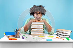 Lack of motivation. Bored african american teen guy sitting at table and leaning on stack of books, blue background