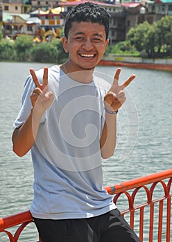 A Ladakhi young guy smiling sitting on safety barrier by lake with making victory hand sign and looking at camera