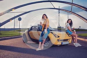 Ladies are smiling, holding hamburgers and beverages in paper cups while sitting on the hood of yellow car cabrio. Fast