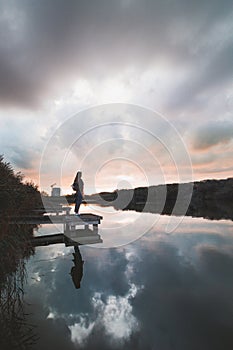 Lady aged 20-25 standing on a wooden pier by a pond watching the sunset over the town of Oostende, western Belgium. Dramatic red-