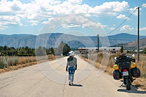 Lady Biker walking down the center of the road, travel motorcycle on the side, summer, active lifestyle, vacation concept Magliano