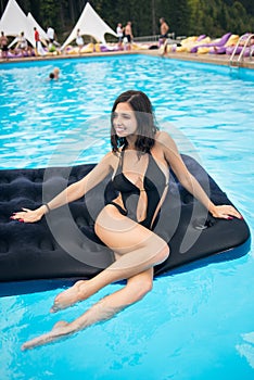 Lady in black bikini sitting on an inflatable mattress in swimming pool and looking away. Blurred background