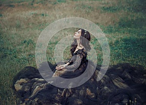 lady in black elegant dress sits on green grass. eyes closed, enjoys nature.
