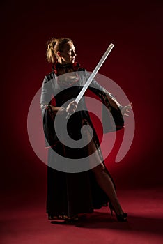 Lady in black gown with a sword standing and posing in studio. Portrait of beautiful elegant woman in evening dress.