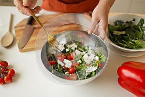 Woman putting cut feta cheese into bowl with tasty salad on table in kitchen and light blur background.