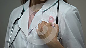 Lady doctor attaching pink ribbon to white medical suit, breast cancer awareness