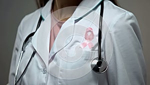 Lady doctor attaching pink ribbon to white medical suit, breast cancer awareness