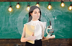 Lady in formal wear on calm face in classroom. Biology concept. Teacher of biology holds book and microscope. Lady